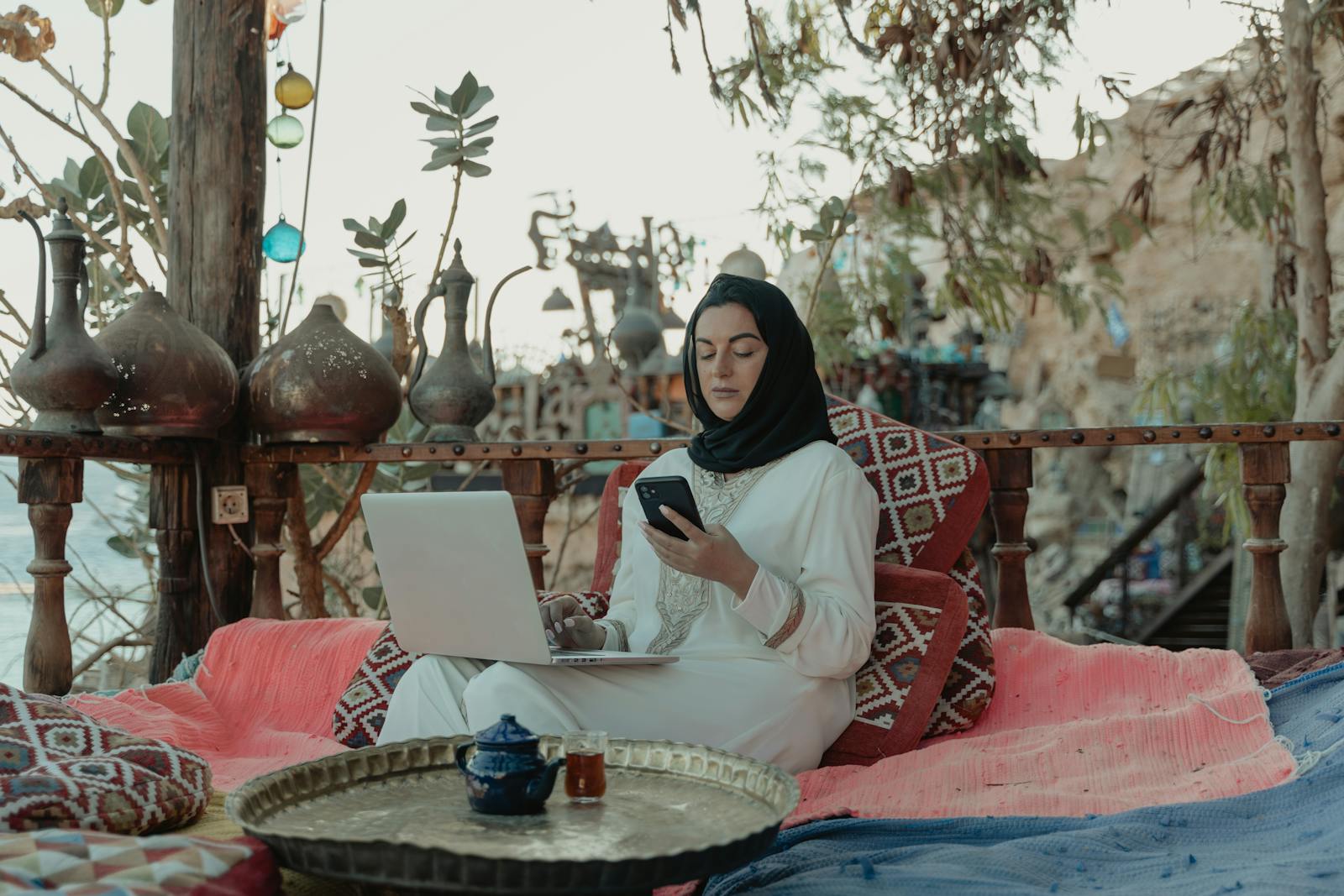 A woman in a hijab working on a laptop and phone in a cozy outdoor setting.